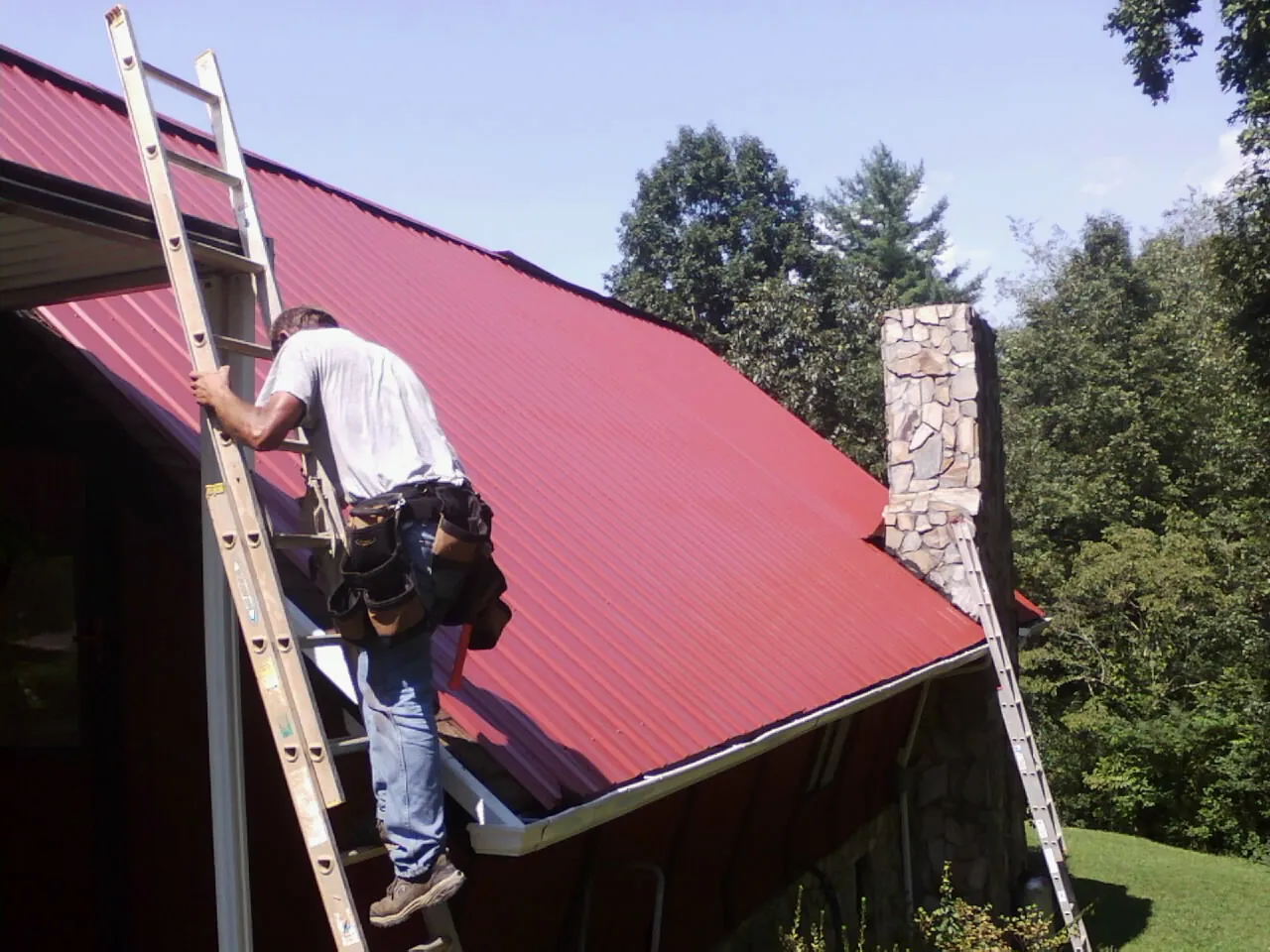 Roofer on a ladder beside a red metal roof and stone chimney — Soffit & Fascia Repair by Roofing Brothers in Mount Pulaski